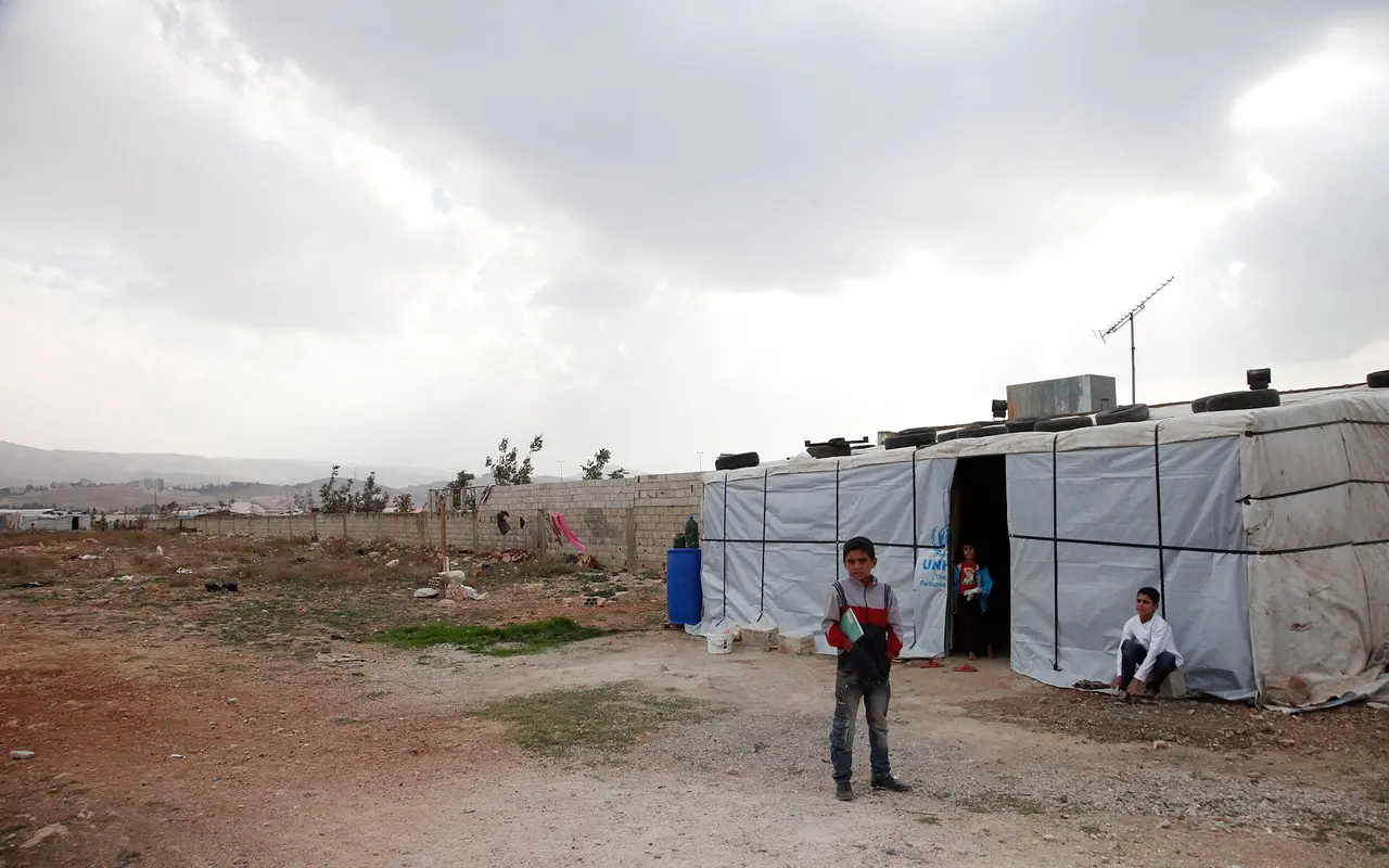 A Syrian boy standing outside a tent shelter made of timber and plastic sheeting in an informal camp