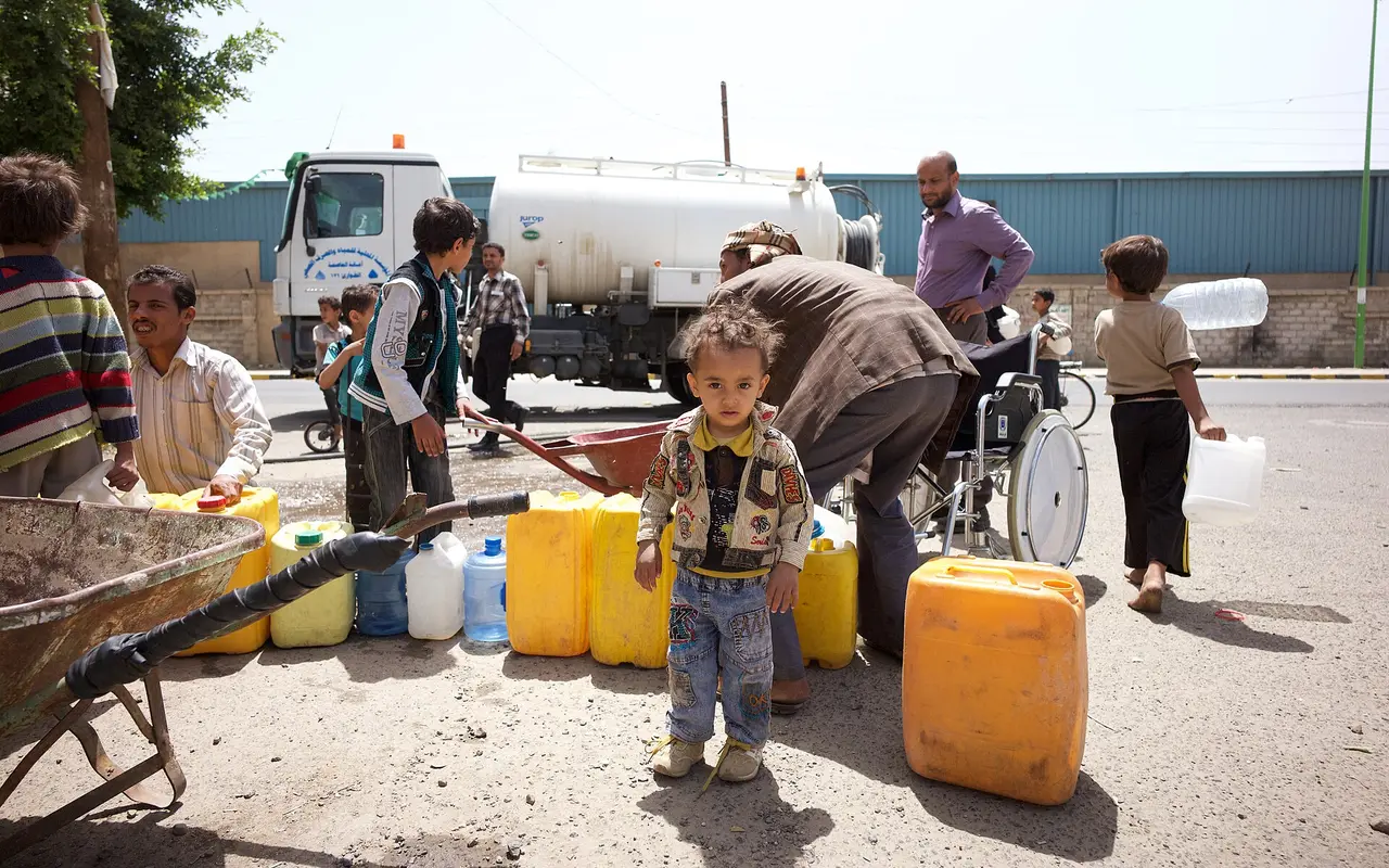 A small child stands among yellow water containers as families collect water from a tanker truck in Sanaa